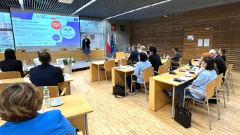 Grupa osób w strojach biznesowych siedzi przy biurkach, naprzeciwko prelegenta prezentującego slajdy w nowoczesnej sali konferencyjnej. W pobliżu prelegenta wyświetlane są flagi i emblematy herbowe. Uczestnicy słuchają uważnie, z notatnikami, napojami i laptopami na biurkach.