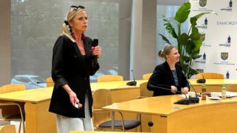 Kobieta w czarnej marynarce stoi trzymając mikrofon, przemawiając w sali konferencyjnej. Inna kobieta siedzi przy biurku z papierami i butelkami. Za nimi stoją drewniane krzesła, roślina i banery z herbem i logo województwa opolskiego.