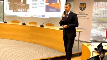 Mężczyzna w garniturze mówi do mikrofonu, stojąc w nowoczesnej sali konferencyjnej. Za nim znajduje się zakrzywiony stół z pustymi krzesłami i dużym ekranem wyświetlającym slajdy. Baner z herbem i tekstem stoi po prawej stronie w pobliżu dużych okien.