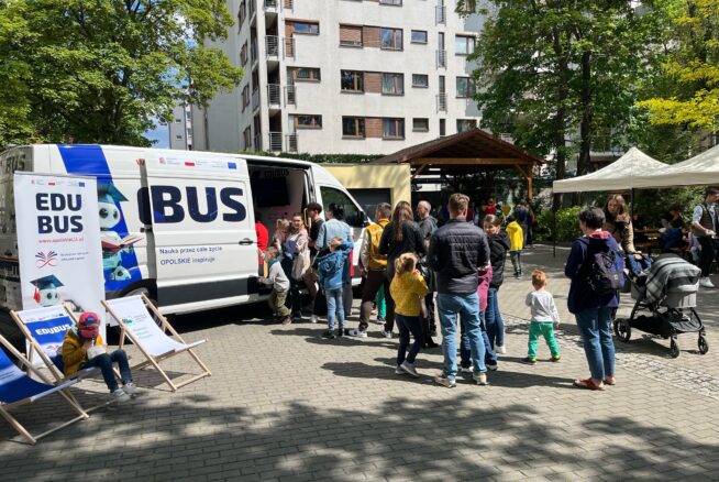 Biała furgonetka EDU BUS jest zaparkowana na utwardzonym terenie w pobliżu budynku mieszkalnego. Wokół furgonetki gromadzą się ludzie, w tym dzieci i dorośli. W pobliżu znajdują się dwa krzesła informacyjne i namioty. Drzewa i światło słoneczne tworzą żywą atmosferę wydarzenia.