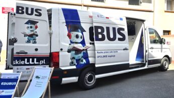 Biała furgonetka z napisem EDUBUS z rysunkowymi robotami trzymającymi książki namalowanymi na bocznych i tylnych drzwiach. Boczne i tylne drzwi furgonetki są otwarte, odsłaniając wnętrze. Na zewnątrz widoczne są znaki i przenośny stojak. Miejscem akcji jest miejska ulica.