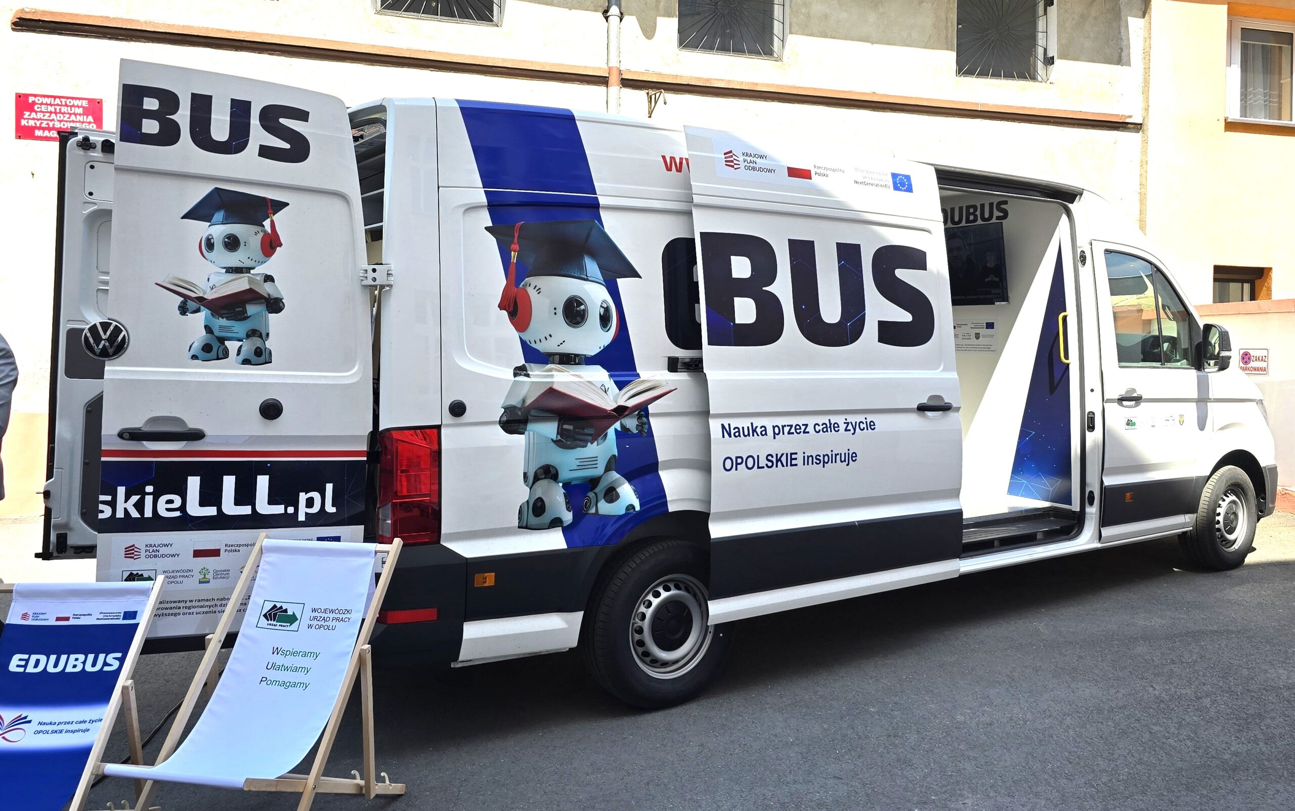 Biała furgonetka z napisem EDUBUS z rysunkowymi robotami trzymającymi książki namalowanymi na bocznych i tylnych drzwiach. Boczne i tylne drzwi furgonetki są otwarte, odsłaniając wnętrze. Na zewnątrz widoczne są znaki i przenośny stojak. Miejscem akcji jest miejska ulica.