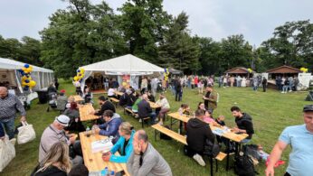 Ludzie siedzą przy drewnianych stołach piknikowych na trawiastym terenie podczas festiwalu na świeżym powietrzu. W tle widać białe namioty i stragany z balonami, a wielu uczestników je, rozmawia lub spaceruje. Wokół rosną drzewa, a niebo jest zachmurzone.