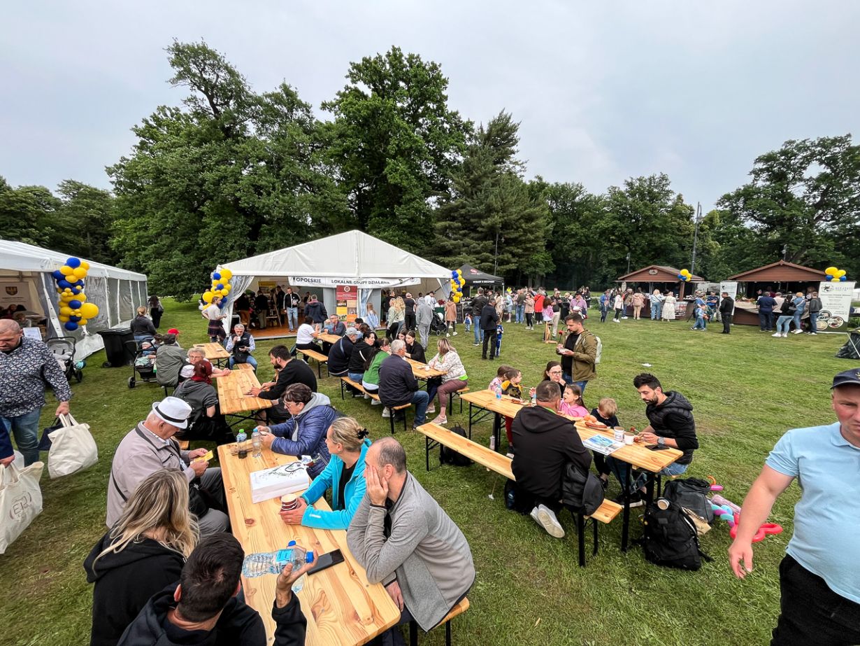 Ludzie siedzą przy drewnianych stołach piknikowych na trawiastym terenie podczas festiwalu na świeżym powietrzu. W tle widać białe namioty i stragany z balonami, a wielu uczestników je, rozmawia lub spaceruje. Wokół rosną drzewa, a niebo jest zachmurzone.