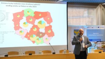 Mężczyzna w garniturze prezentuje się w sali konferencyjnej z dużym ekranem pokazującym mapę Polski. Mapa wyświetla dane dotyczące zmiany liczby ludności w 2023 r. za pomocą zielonych i czerwonych kolorów oraz wykresów kołowych, z tekstem i wykresami w języku polskim.
