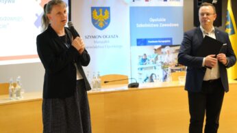 Kobieta w czarnej marynarce mówi do mikrofonu, a mężczyzna w garniturze trzyma obok niej teczkę. Stoją w sali konferencyjnej z ekranem projektora, banerami województwa opolskiego i żółtą flagą w tle. Na stole stoją butelki wody.