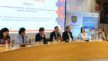 Sześciu profesjonalistów, czterech mężczyzn i dwie kobiety, siedzi przy zakrzywionym stole z mikrofonami w sali konferencyjnej. Ekran projekcyjny za nimi wyświetla polski tekst na temat biznesu, edukacji i współpracy. Branding i logo są widoczne, a jeden mężczyzna przemawia.