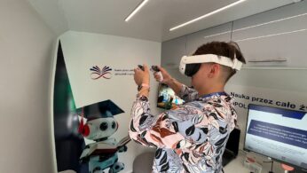 Osoba nosząca biały headset VR i trzymająca kontrolery stoi w nowoczesnym pokoju z białymi ścianami. Za nimi znajduje się plakat przedstawiający robota i tekst w języku polskim. Monitor komputera i klawiatura są widoczne na blacie po prawej stronie.