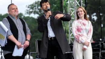 Trzy osoby stoją na scenie na świeżym powietrzu. Mężczyzna w centrum, trzymający mikrofon, gestykuluje podczas mówienia. Ma na sobie ciemny płaszcz. Po jego prawej stronie stoi uśmiechnięta kobieta w różowej bluzce w kwiaty ze splecionymi dłońmi. Inny mężczyzna stoi po lewej stronie, trzymając dokumenty.