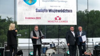 Cztery osoby stoją na scenie pod gołym niebem. Kobieta z mikrofonem stoi przed trzema mężczyznami; jeden trzyma schowek, a dwóch trzyma dokumenty. Za nimi znajduje się duży baner w języku polskim o regionalnym wydarzeniu, z instrumentami muzycznymi i drzewami widocznymi w tle.