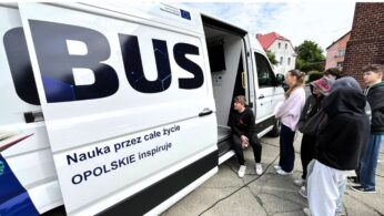 Grupa młodych ludzi stoi przed białą furgonetką z dużym napisem BUS na boku. Drzwi furgonetki są otwarte, a przy wejściu kuca jedna osoba. Tekst na furgonetce brzmi: Nauka przez całe życie OPOLSKIE inspiruje. W tle widoczne są budynki i drzewa.