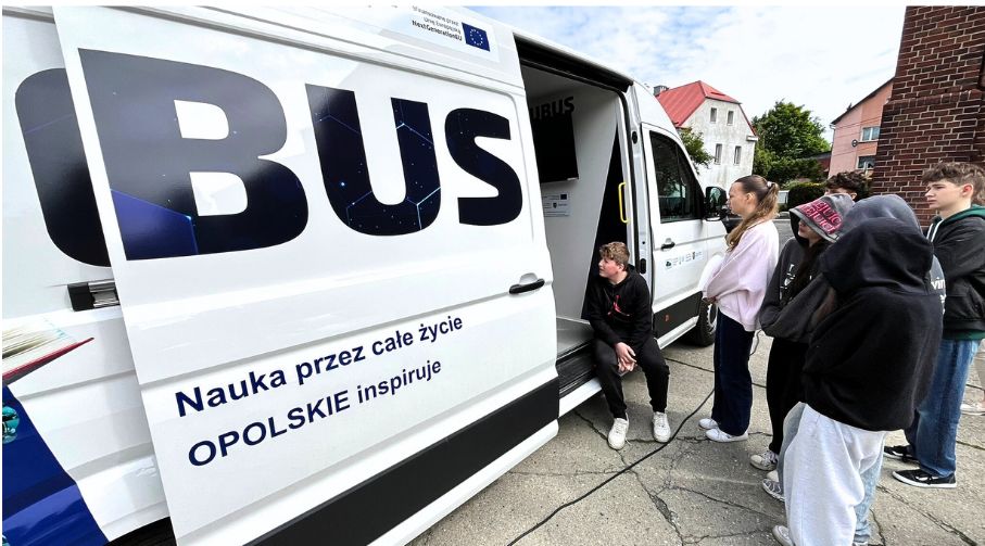 Grupa młodych ludzi stoi przed białą furgonetką z dużym napisem BUS na boku. Drzwi furgonetki są otwarte, a przy wejściu kuca jedna osoba. Tekst na furgonetce brzmi: Nauka przez całe życie OPOLSKIE inspiruje. W tle widoczne są budynki i drzewa.