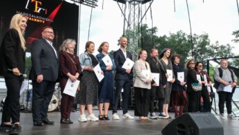 Grupa dwunastu osób stoi na scenie pod gołym niebem, uśmiechając się i trzymając certyfikaty. Za nimi znajduje się duży ekran wyświetlający 