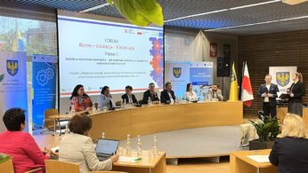 Panel składający się z dziewięciu osób siedzi przy zakrzywionym stole w sali konferencyjnej, twarzą do publiczności. Duży ekran za nimi wyświetla prezentację w języku polskim. Dwie stojące kobiety zwracają się do sali. Banery, flagi i członkowie publiczności są widoczni w całej nowoczesnej przestrzeni.