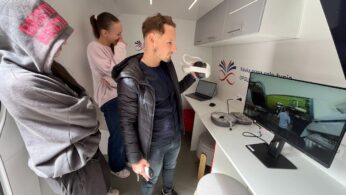 Trzy osoby znajdują się w nowoczesnym białym pokoju z szafkami. Jeden mężczyzna trzyma zestaw VR i patrzy na monitor komputera pokazujący scenę na zewnątrz. Dwie kobiety stoją w pobliżu, jedna w bluzie z kapturem z rękami w kieszeniach, druga z ręką przy ustach.