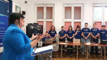 Kobieta w niebieskiej marynarce przemawia z podium do grupy siedmiu młodych ludzi w dopasowanych niebieskich koszulach, stojących za stołami z laptopami i urządzeniami elektronicznymi, w klasie z dużymi oknami i plakatami na ścianie.