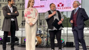 Cztery osoby stoją na scenie na świeżym powietrzu z instrumentami muzycznymi za nimi. Mężczyzna trzymający mikrofon mówi, podczas gdy inni słuchają. Kobieta w kwiecistej bluzce stoi między dwoma mężczyznami w garniturach. Nad nimi wyświetlany jest duży napis wydarzenia w języku polskim.