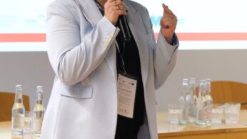 Osoba z ciemnymi włosami do ramion, ubrana w jasnoniebieską marynarkę i czarne spodnie, mówi do mikrofonu podczas konferencji. Stoją przed drewnianym stołem z butelkowaną wodą i dużym ekranem wyświetlającym tekst i kolorowe abstrakcyjne logo.