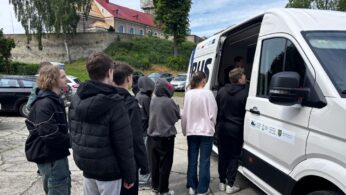 Grupa nastolatków, ubranych w luźne kurtki i bluzy z kapturem, stoi w kolejce przed białą furgonetką z logo na drzwiach. Furgonetka jest zaparkowana na parkingu, z zaparkowanymi samochodami i kościołem z czerwonym dachem i dzwonnicą w tle. Widoczne są drzewa i zachmurzone niebo.