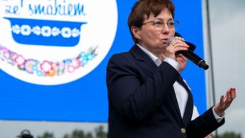 Kobieta w ciemnym garniturze i okularach mówi do mikrofonu na scenie. Gestykuluje jedną ręką. Za nią znajduje się niebieski ekran z logo 