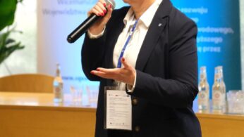 Osoba w ciemnym garniturze, białej koszuli i okularach mówi do mikrofonu, gestykulując ręką. Za nimi znajdują się banery z niebieskimi i żółtymi wzorami, butelkowana woda na stole i zielone rośliny, co sugeruje formalne wydarzenie lub konferencję.