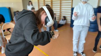Dwoje młodych ludzi noszących zestawy VR stoi w pokoju z matami gimnastycznymi i drewnianymi drążkami na ścianach. Dziewczyna z przodu pochyla się do przodu, trzymając kontrolery VR, wyglądając na zaangażowaną. Tło pokazuje więcej osób siedzących i rozmytych, obserwujących aktywność.