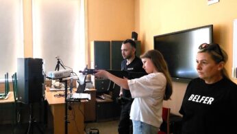 Dziewczynka celuje z symulatora karabinu w klasie w asyście mężczyzny w koszulce Policji. Kobieta w koszulce Belfer obserwuje. Sprzęt komputerowy, projektor i duży monitor są widoczne wśród biurek i krzeseł w klasie.