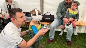 Mężczyzna klęczy obok małego dziecka noszącego zestaw VR i trzymającego kontrolery. W pobliżu inny mężczyzna siedzi na ławce, trzymając dziecko z pomalowaną twarzą. Za nimi siedzi kobieta. Wszyscy znajdują się w namiocie, a podłogę stanowi trawa.
