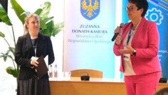 Dwie kobiety stoją przed drewnianym stołem i transparentami. Kobieta po lewej ma na sobie ciemną kurtkę i sukienkę w kropki, trzyma teczkę. Kobieta po prawej ma na sobie jasnoróżowy garnitur i okulary, mówi do mikrofonu. Za nimi widoczna jest duża roślina.