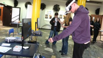 Młoda osoba nosząca gogle VR stoi przed ekranem, wchodząc w interakcję z wirtualnym środowiskiem za pomocą gestów dłoni. Kilka osób w tle obserwuje lub korzysta z telefonów w przestronnym, nowoczesnym pokoju z żółtymi filarami i plakatami na ścianie.