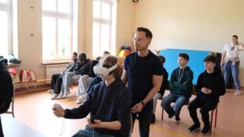 Grupa uczniów siedzi w klasie z dużymi oknami. Jeden uczeń na pierwszym planie nosi zestaw VR i trzyma kontrolery, podczas gdy dorosły stoi obok niego i nadzoruje. Kilku innych uczniów obserwuje, niektórzy wydają się zaangażowani, a inni zrelaksowani.