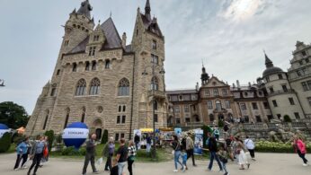 Duży, ozdobny zamek ze spiczastymi wieżami i łukowatymi oknami wznosi się za ruchliwym placem wypełnionym spacerującymi ludźmi, niektórymi z wózkami i dziećmi. Wokół porozrzucane są kolorowe stoiska i niebieskie balony, a niebo jest w większości zachmurzone.