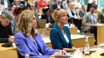 Dwie kobiety siedzą uważnie w jasnej, nowoczesnej sali konferencyjnej wśród wielu uczestników. Obie mają na sobie marynarki, a na biurku przed nimi leżą notatniki i butelki z wodą. Tło przedstawia tłum słuchający, z widocznymi laptopami i papierami.