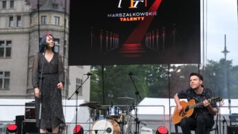 Kobieta śpiewa do mikrofonu, a mężczyzna gra na gitarze akustycznej na scenie plenerowej. Za nimi zestaw perkusyjny i duży ekran wyświetlający tekst 