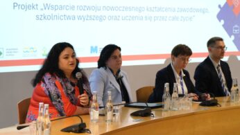 Cztery osoby siedzą przy stole konferencyjnym z mikrofonami i butelkowaną wodą, uczestnicząc w panelu. Za nimi slajd prezentacji w języku polskim omawia projekt wspierający nowoczesne szkolnictwo zawodowe i wyższe. Jedna z kobiet mówi do mikrofonu.