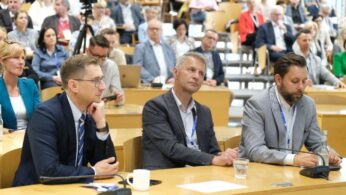 Duża grupa ludzi siedzi w sali wykładowej, uważnie słuchając. Trzech mężczyzn w garniturach siedzi przy przednim stole z mikrofonami, filiżanką kawy i szklanką wody. Większość uczestników zwrócona jest twarzami do przodu, co sugeruje, że jest to formalna konferencja lub seminarium.