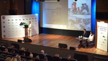 Kobieta prezentuje na scenie dla publiczności w audytorium. Stoi przy ekranie wyświetlającym slajdy z tekstem i obrazami. Dwóch siedzących panelistów stoi twarzą do publiczności. Banery z logo i tekstem są umieszczone po obu stronach sceny.