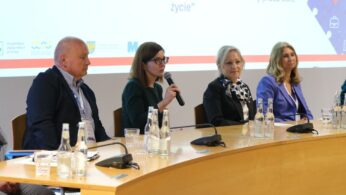 Pięć osób siedzi przy zakrzywionym stole konferencyjnym z mikrofonami i butelkami wody. Kobieta w okularach mówi do mikrofonu. Za nimi znajduje się ekran wyświetlający polski tekst i kolorową grafikę. Grupa wydaje się być zaangażowana w dyskusję panelową lub prezentację.