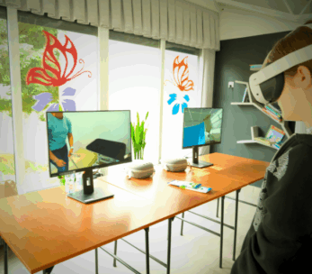 Osoba nosząca gogle VR stoi w pobliżu drewnianych stołów z dwoma monitorami komputerowymi wyświetlającymi wirtualne środowiska. Pokój ma duże okna z naklejkami motyli, rośliny doniczkowe i półki z książkami i dekoracjami w tle.