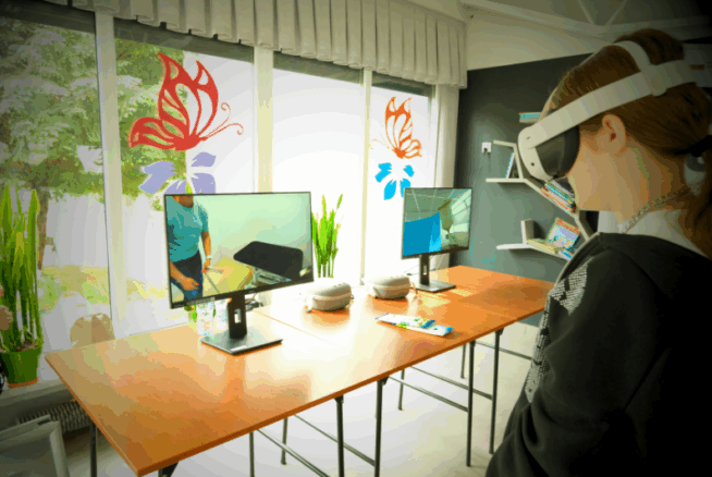 Osoba nosząca gogle VR stoi w pobliżu drewnianych stołów z dwoma monitorami komputerowymi wyświetlającymi wirtualne środowiska. Pokój ma duże okna z naklejkami motyli, rośliny doniczkowe i półki z książkami i dekoracjami w tle.