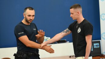 Policjant w mundurze demonstruje młodemu mężczyźnie w czarnej koszulce technikę samoobrony w klasie z niebieską ścianą i białą tablicą. Funkcjonariusz wyjaśnia, podczas gdy młody człowiek podąża za nim.
