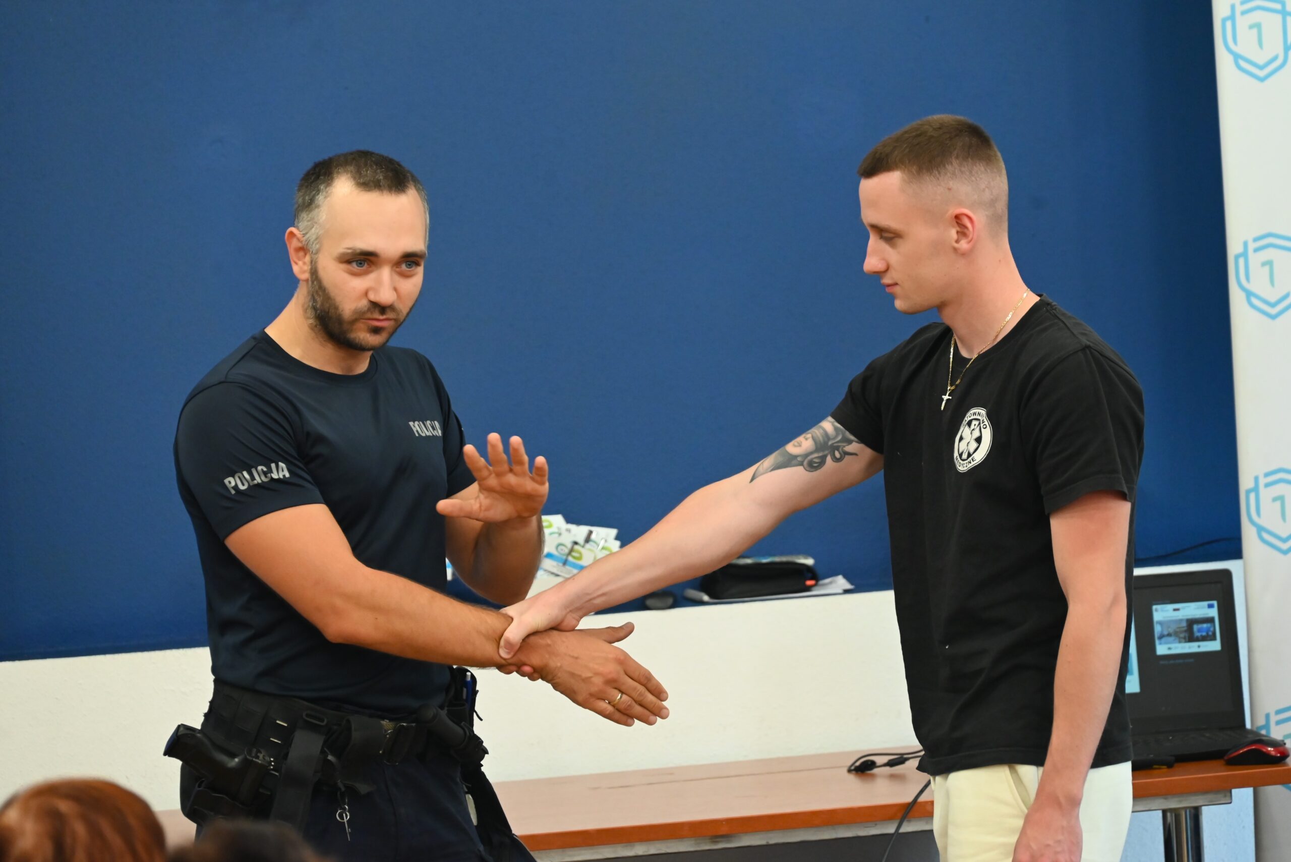 Policjant w mundurze demonstruje młodemu mężczyźnie w czarnej koszulce technikę samoobrony w klasie z niebieską ścianą i białą tablicą. Funkcjonariusz wyjaśnia, podczas gdy młody człowiek podąża za nim.