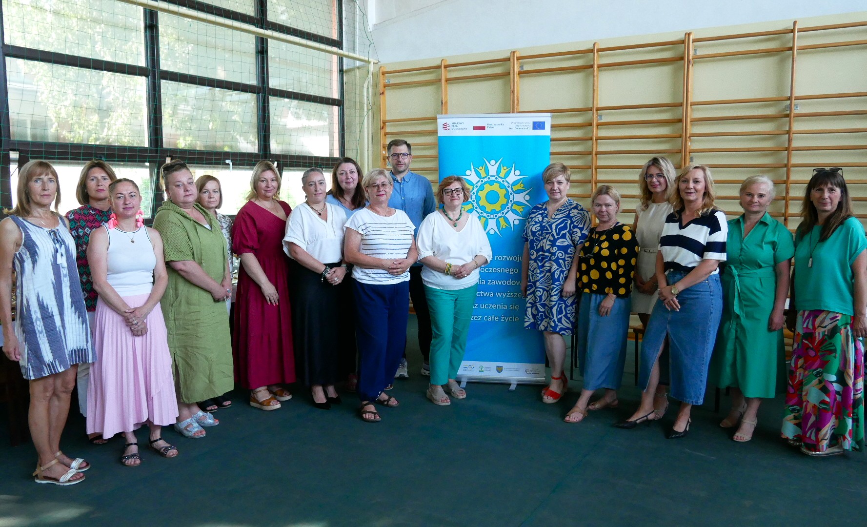Grupa 15 dorosłych osób, w większości kobiet, stoi w pomieszczeniu przed ścianą z drewnianymi prętami i kolorowym banerem projektu. Ubrani są w różne codzienne i półformalne stroje, pozują do grupowego zdjęcia i uśmiechają się do kamery.