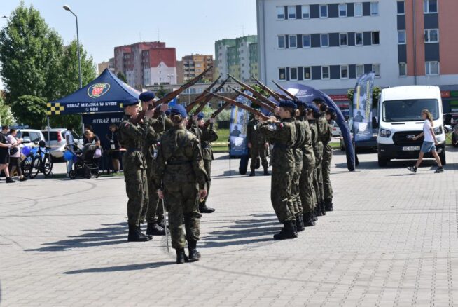 Grupa umundurowanych żołnierzy stoi w dwóch liniach na wybrukowanym placu, unosząc karabiny w uroczystym łuku. Za nimi znajdują się namioty, zaparkowana furgonetka i nowoczesne budynki mieszkalne. Wydarzeniu przyglądają się widzowie, niektórzy na siedząco, z rowerami w pobliżu.