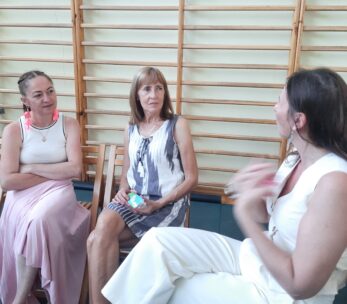 Trzy kobiety siedzą i rozmawiają pod drewnianą ścianą sali gimnastycznej. Dwie kobiety, jedna w różowej spódnicy i jedna we wzorzystej sukience, słuchają uważnie. Trzecia kobieta, ubrana na biało, gestykuluje ręką podczas mówienia. Wszystkie wydają się być zaangażowane w rozmowę.