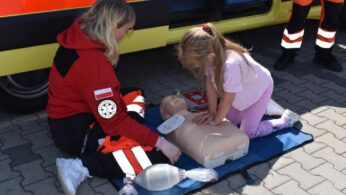 Ratownik medyczny prowadzi młodą dziewczynę podczas wykonywania uciśnięć klatki piersiowej na manekinie treningowym CPR na zewnątrz, w pobliżu karetki pogotowia. Sprzęt medyczny, w tym worek resuscytacyjny, leży w pobliżu na macie. Oboje skupiają się na demonstracji.