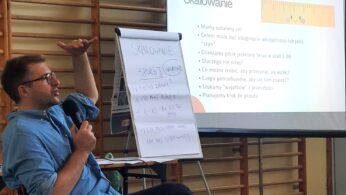 Mężczyzna w okularach trzyma mikrofon i gestykuluje siedząc. Obok niego znajduje się flipchart z odręcznymi notatkami. Za nim wyświetlany jest slajd zatytułowany 
