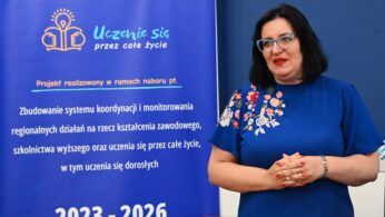 Kobieta w okularach i ciemnych włosach, ubrana w niebieską sukienkę z kolorowym kwiatowym haftem, stoi obok niebieskiego baneru w języku polskim na temat projektu uczenia się przez całe życie dla dorosłych, trwającego od 2023 do 2026 roku. Kobieta uśmiecha się i jest zwrócona lekko w prawo.