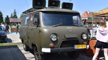 Na parkingu stoi zabytkowa oliwkowozielona furgonetka z trzema dużymi głośnikami na górze. Furgonetka ma pudełkowaty kształt, okrągłe reflektory i żółtą tablicę rejestracyjną z napisem ONY 12. W tle widoczni są ludzie i domy w słoneczny dzień.