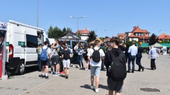 Grupa ludzi, głównie młodych, gromadzi się w pobliżu białej furgonetki i namiotów na imprezie plenerowej w słoneczny dzień. Niektórzy stoją w kolejce, inni spacerują. Wokół znajdują się budynki z czerwonymi dachami, zielone drzewa i czyste, błękitne niebo.